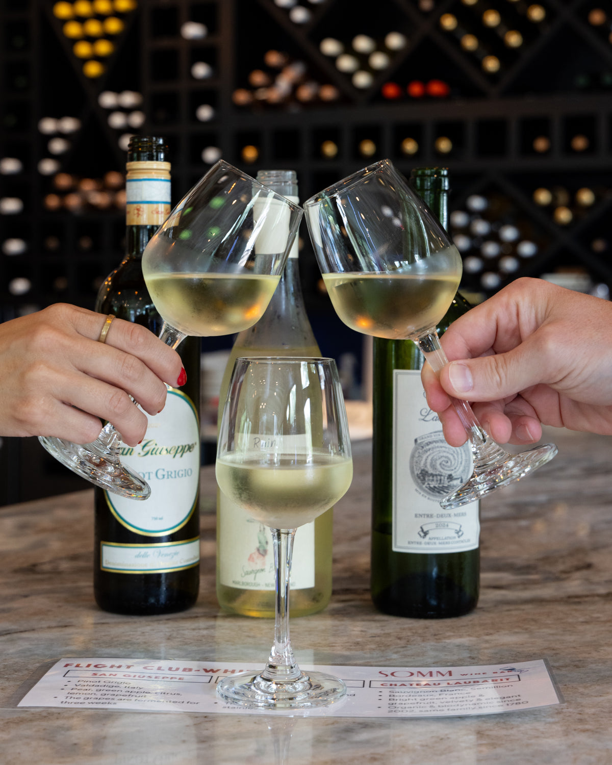 Three wine bottles and three stemmed wine glasses filled with white wine are arranged on a table, with two hands holding the glasses in a clinking toast pose. A wine bottle label reading 'G.