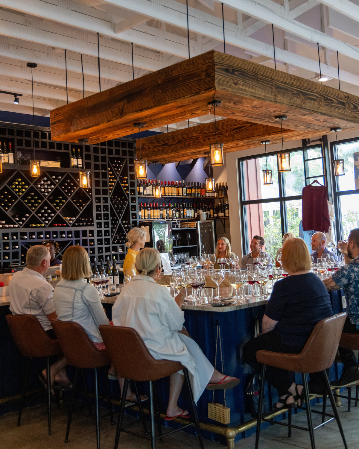 A group of people seated at a long bar table in a restaurant or wine bar, with wine glasses, bottles, shelving, and large wooden ceiling beams visible in the indoor setting.
